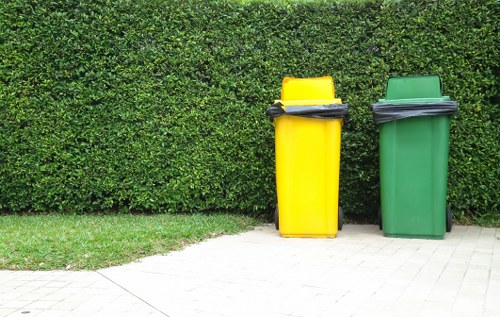 In the foreground, two upright wheelie bins are positioned on a paved pathway near a well-maintained green hedge. The yellow bin on the left lacks a lid and appears filled with black rubbish bags stacked on top, while the green bin on the right has a closed black lid. The hedge in the background is dense and neatly trimmed, providing a natural backdrop that suggests a residential garden or driveway setting typical in Hounslow. The pathway's light-colored paving contrasts with the vibrant greenery, emphasizing the bins’ bright colors. This scene may reflect typical rubbish collection points in the area, managed or serviced by Garden Clearance Hounslow, a company specialising in waste removal and garden clearance services around the local postcode. The lighting is natural, with no shadows indicating an overcast day, contributing to a clear, neutral portrayal of waste disposal in a suburban environment.