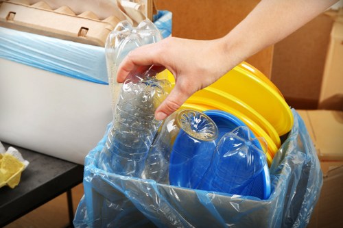 A person's hand is seen holding a clear plastic bottle filled with liquid, hovering above a blue recycling bin lined with a transparent plastic bag. Inside the bin, there are multiple brightly coloured plastic objects, including yellow and blue lids or containers, which appear to be household or garden-related items. The bin is situated on a surface, possibly in a residential or garden area in Hounslow, with a background that suggests an indoor or sheltered outdoor space. The scene captures the preparation for rubbish collection or recycling, reflecting typical waste management activities undertaken by companies like Garden Clearance Hounslow. The environment shown is tidy, with the focus on the safe handling and disposal of plastic waste material, indicative of local rubbish removal or recycling services aimed at clearing garden or household clutter in the Hounslow area.