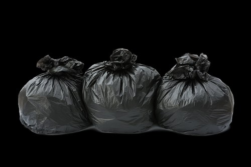 Three black rubbish bags made of thick plastic, filled with waste, are positioned side by side on a dark surface against a black background. The bags are tied at the top with gathered plastic and appear to contain mixed refuse, possibly garden or household waste. The bags have a glossy finish with visible creases and folds, suggesting they are loosely filled. They are aligned horizontally across the middle of the image, with slight variations in size and shape, indicating different amounts of contents inside. The lighting casts subtle reflections on the plastic surface, emphasizing their texture and the volume of the waste inside. This image visually represents rubbish collection or removal services, relevant to providers like Garden Clearance Hounslow who handle waste disposal, particularly in residential or garden settings within the Hounslow area. The dark background isolates the bags, drawing clear focus on the waste containment solution typical of rubbish removal scenarios in London or nearby boroughs.